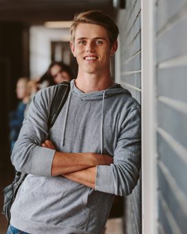 Portrait of handsome young university student standing and leaning to a wall in college corridor with other students at the back. Caucasian male student in high school campus.